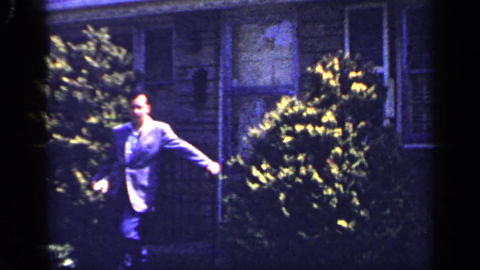 In the 1965 Long Island home-movie frame, the suited man continues across the yard with house windows and plantings behind him.