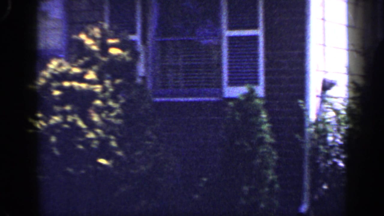 Long Island house exterior in 1965 with white-trimmed windows, siding, shrubs, and a bright side wall after the man leaves frame.