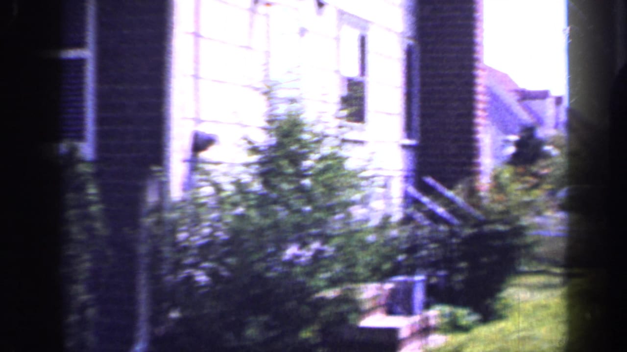 1965 Long Island home-movie frame showing a suburban house exterior, front shrubs, steps, lawn, and heavy film edge shadows.