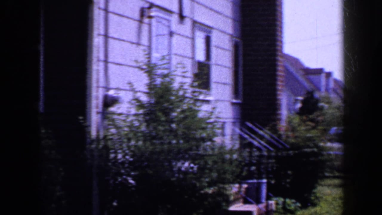1965 Long Island suburban houses with light siding, brick trim, shrubs, steps, and lawn in a soft home-movie frame.