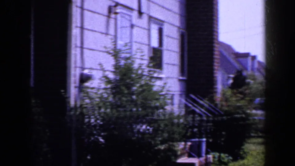 1965 Long Island suburban houses with light siding, brick trim, shrubs, steps, and lawn in a soft home-movie frame.