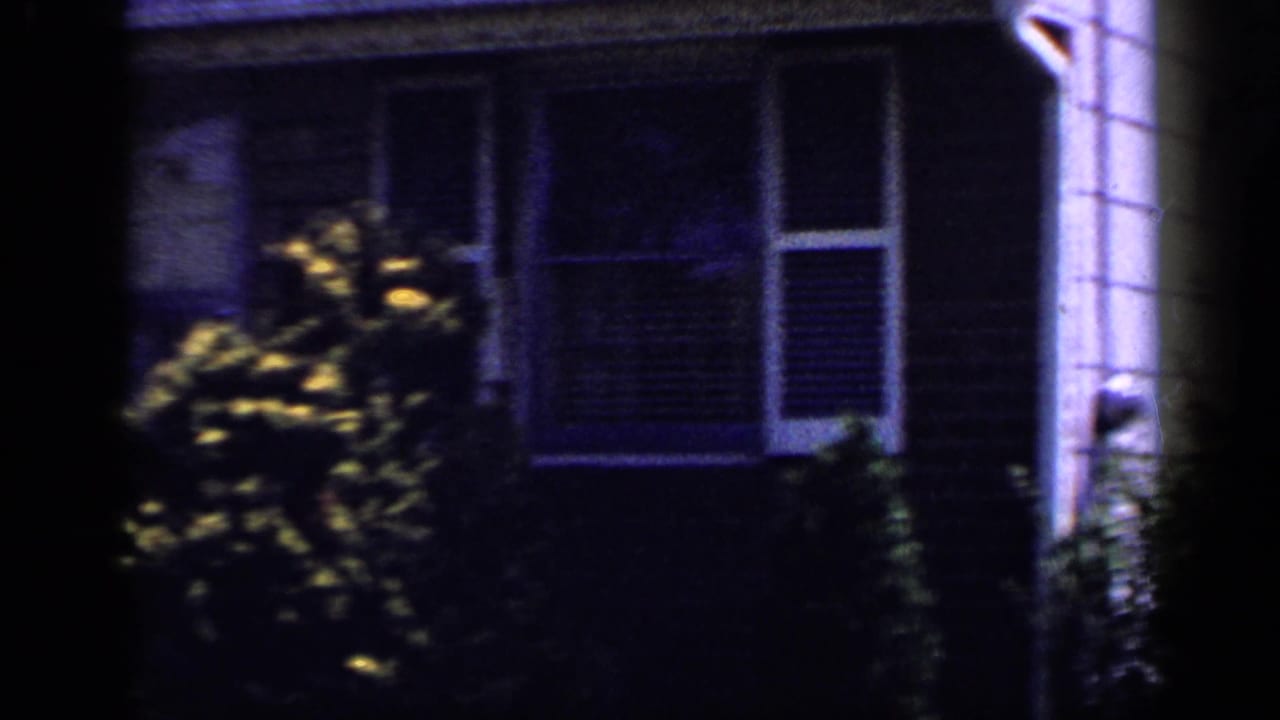 Shaded 1965 Long Island home-movie view of a suburban house facade with bushes, windows, siding, and adjacent house wall.