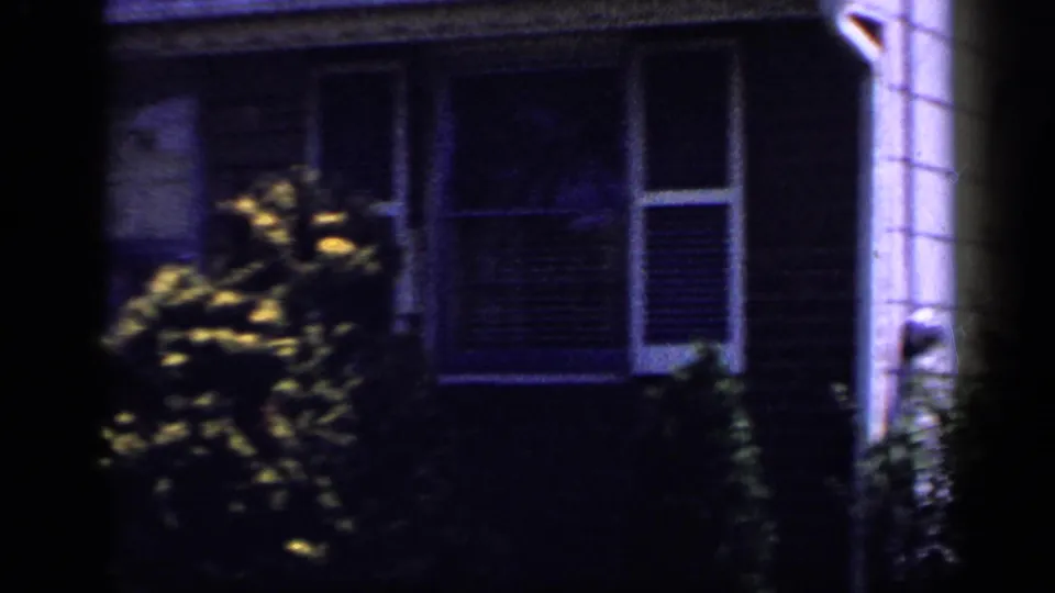 Shaded 1965 Long Island home-movie view of a suburban house facade with bushes, windows, siding, and adjacent house wall.