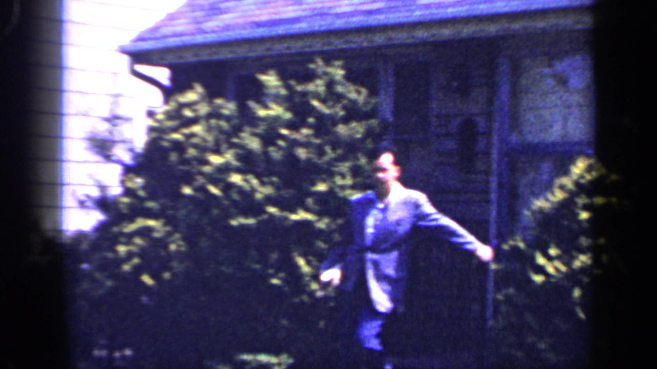 1965 Long Island home-movie frame with a suited man walking on a front lawn near shrubs, porch steps, and a house exterior.