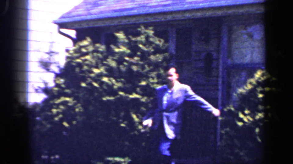 1965 Long Island home-movie frame with a suited man walking on a front lawn near shrubs, porch steps, and a house exterior.
