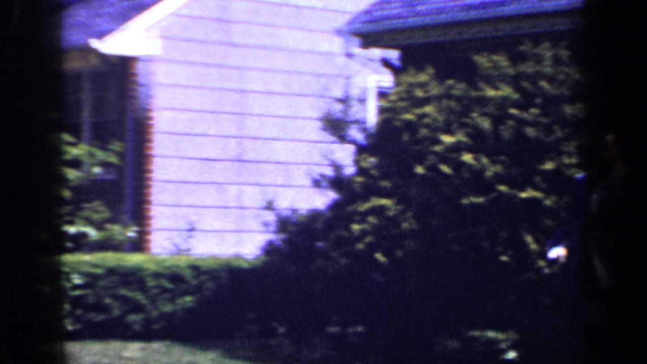 1965 Long Island home-movie frame showing a light-sided suburban house partly obscured by shrubs and hedge-lined lawn.