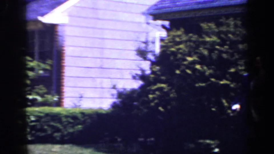 1965 Long Island home-movie frame showing a light-sided suburban house partly obscured by shrubs and hedge-lined lawn.