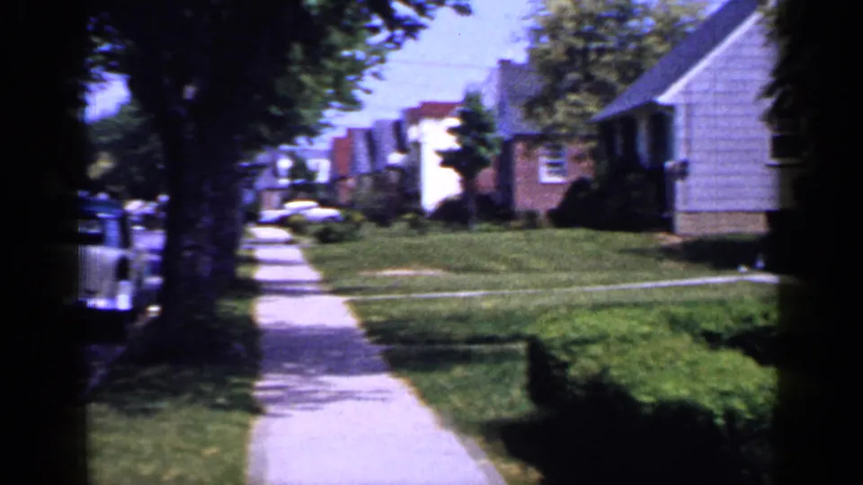 1965 Long Island street-level home-movie view down a concrete sidewalk with houses, lawns, trees, hedges, and a parked car edge.