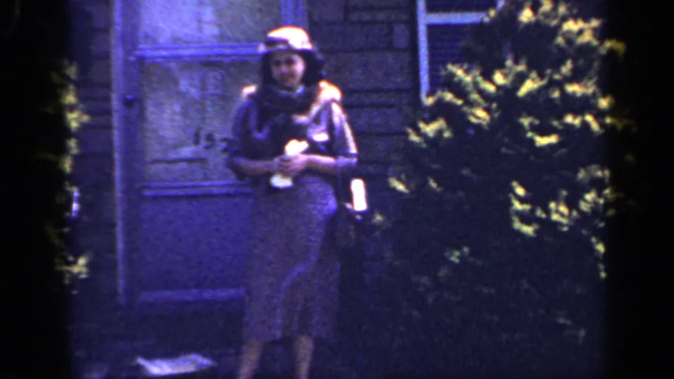 1965 Long Island home-movie frame showing an adult woman in a dress and hat standing outside near a doorway, shrubs, and house exterior.
