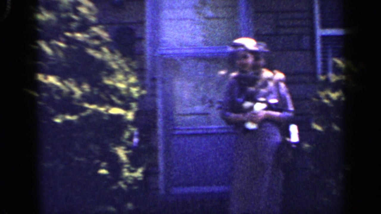 Woman in a hat and dark dress stands outside a house near shrubs.