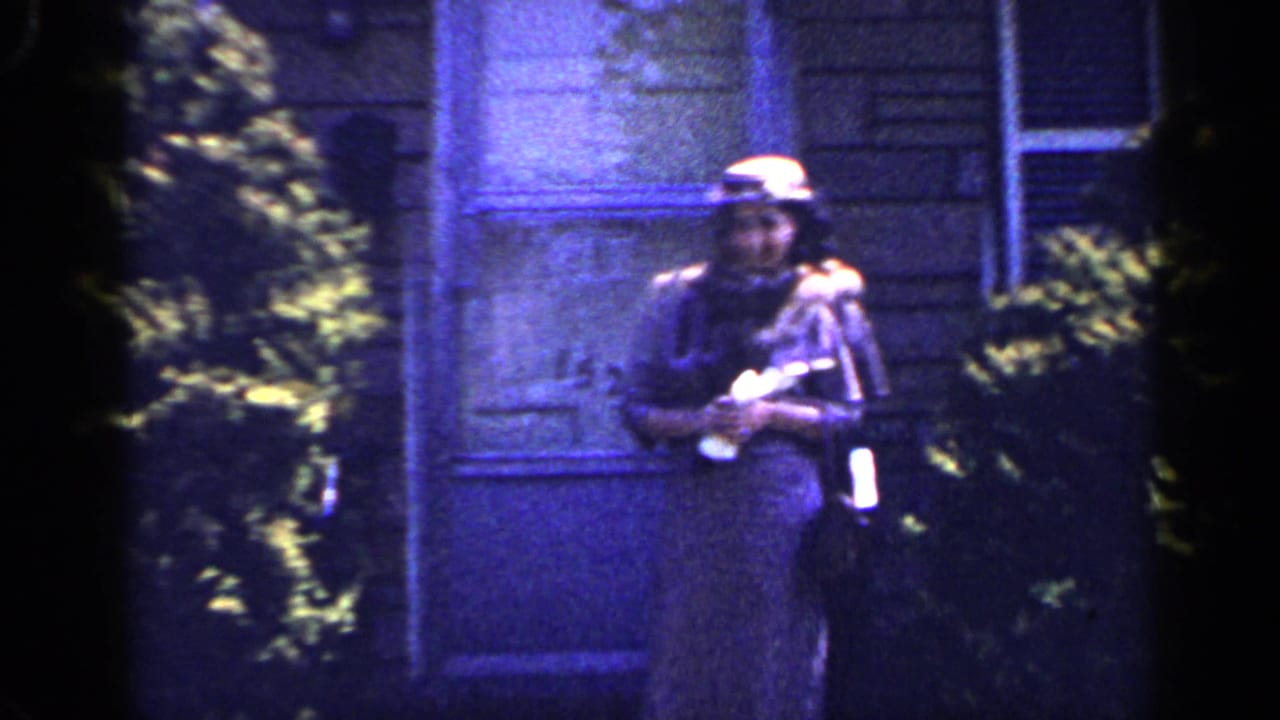 In a blue-tinted 8mm frame, the woman remains near the house exterior, facing the camera with hat, scarf and dress visible.