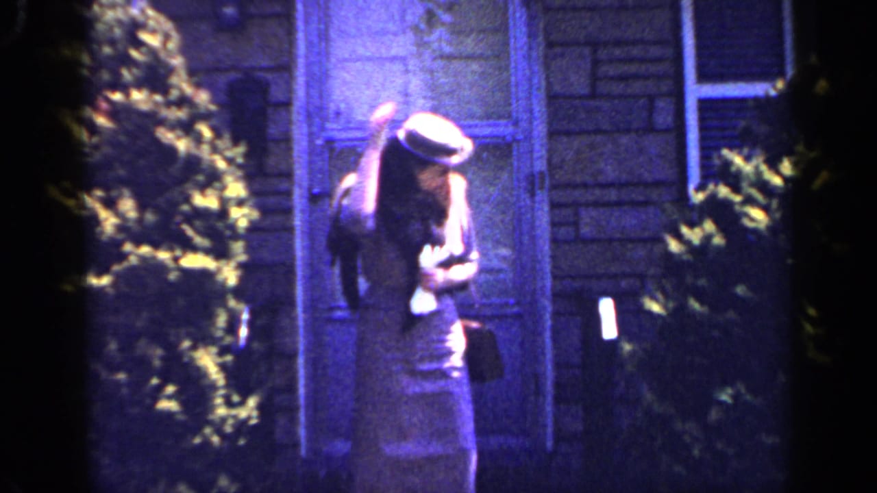 The woman lowers her head and adjusts her brimmed hat near a window and shrubs in the 1965 Long Island home-movie scene.
