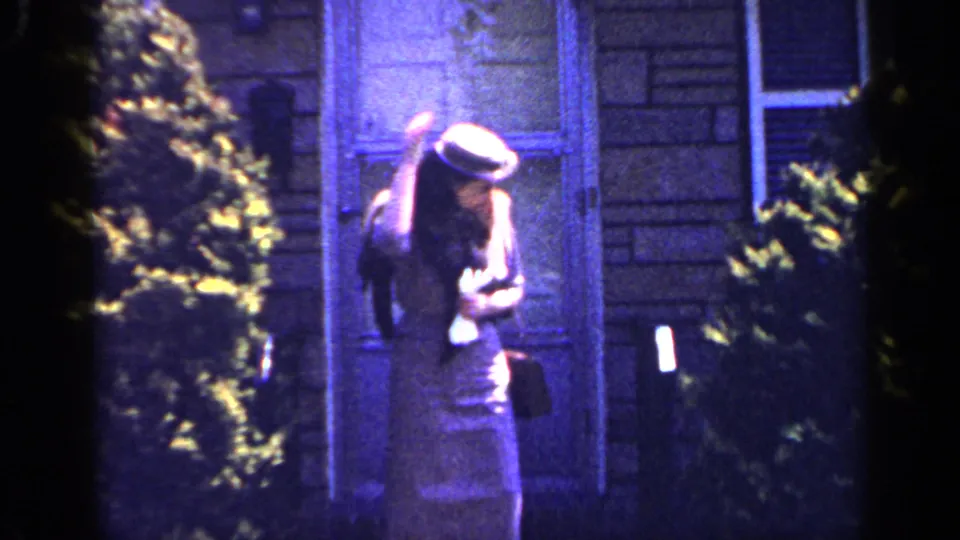 The woman lowers her head and adjusts her brimmed hat near a window and shrubs in the 1965 Long Island home-movie scene.
