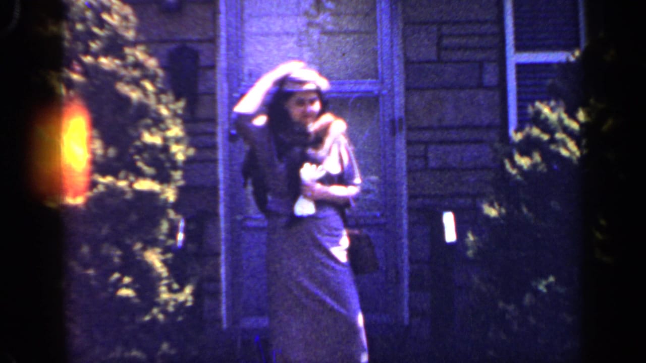 In 1965 Long Island home-movie footage, a woman in a long dress holds her wide-brimmed hat near a house exterior.