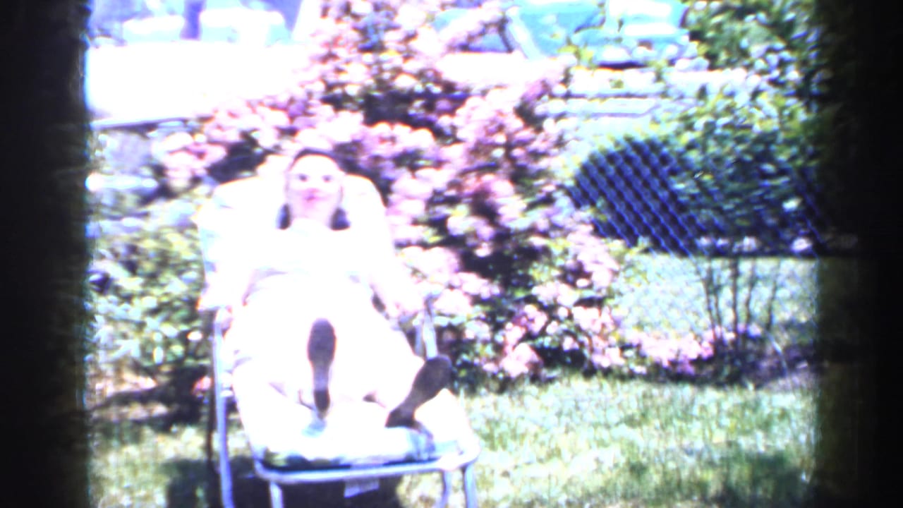 A woman reclines in a metal lawn chair in a sunlit Long Island backyard with flowering shrubs and a chain-link fence.