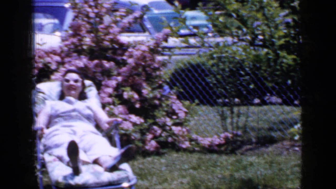 A soft 8mm frame shows the woman reclining in the backyard chair beside pink shrubs and a chain-link fence.