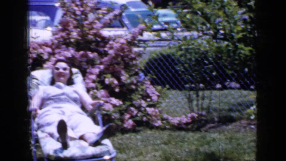A soft 8mm frame shows the woman reclining in the backyard chair beside pink shrubs and a chain-link fence.
