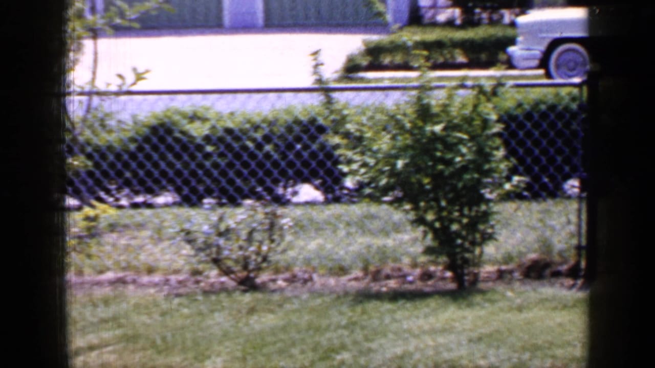 A mid-1960s Long Island street view shows a pale sedan passing beyond a fenced suburban yard.