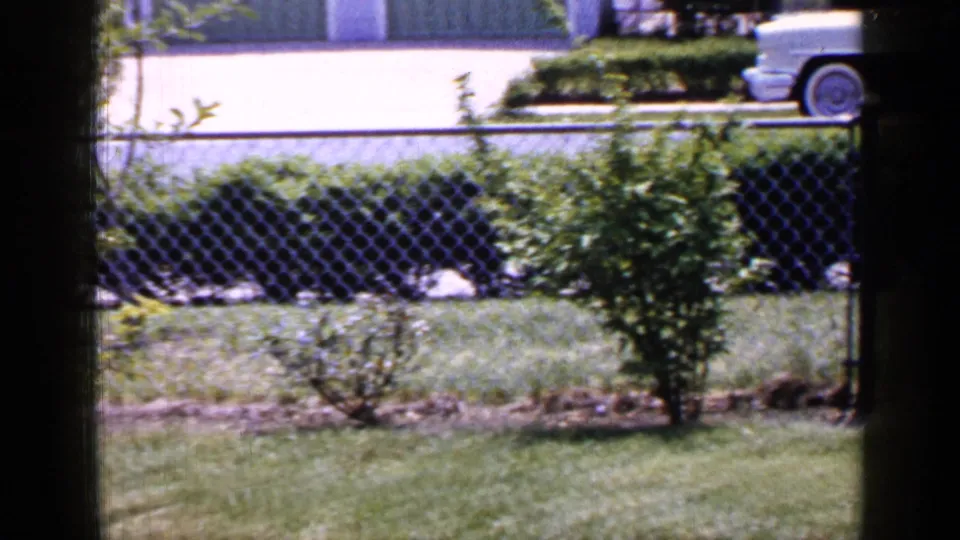 A mid-1960s Long Island street view shows a pale sedan passing beyond a fenced suburban yard.