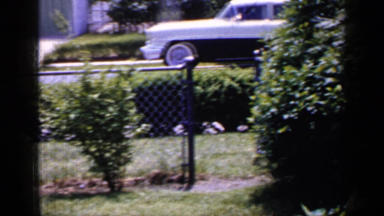 The home-movie view holds on a fenced Long Island yard with shrubs and a light sedan partly visible across the street.
