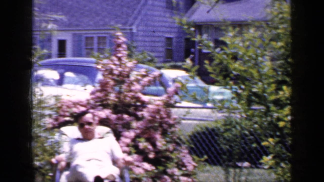 A 1965 Long Island suburban yard frame shows a man seated outdoors near blooming shrubs, with mid-century cars and a shingled residence in the background.