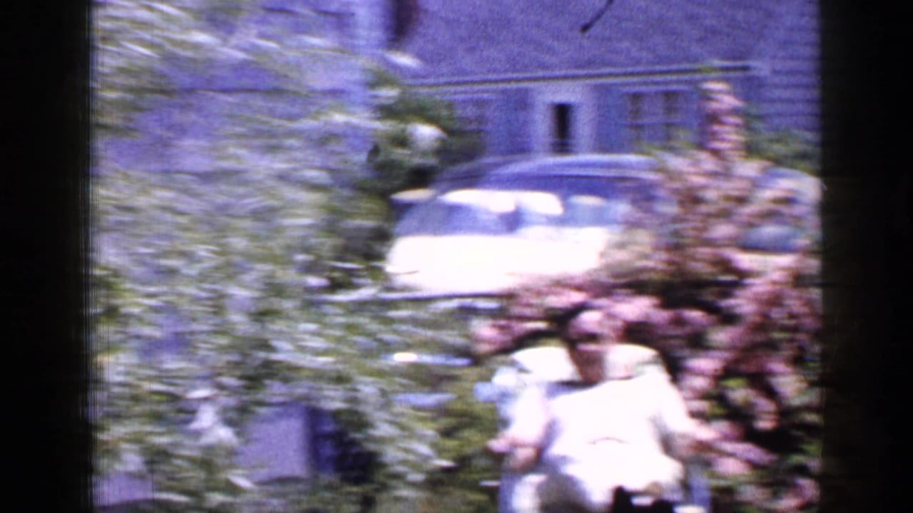 In 1965 Long Island home-movie footage, a man reclines in a backyard near leafy plants, with a light-colored car and residential building visible behind him.