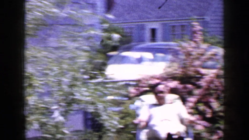In 1965 Long Island home-movie footage, a man reclines in a backyard near leafy plants, with a light-colored car and residential building visible behind him.