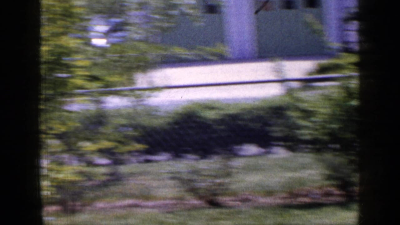 A handheld 1965 Long Island backyard view shows lawn, shrubs, fencing, and nearby residential structures, with the frame blurred by motion and soft focus.