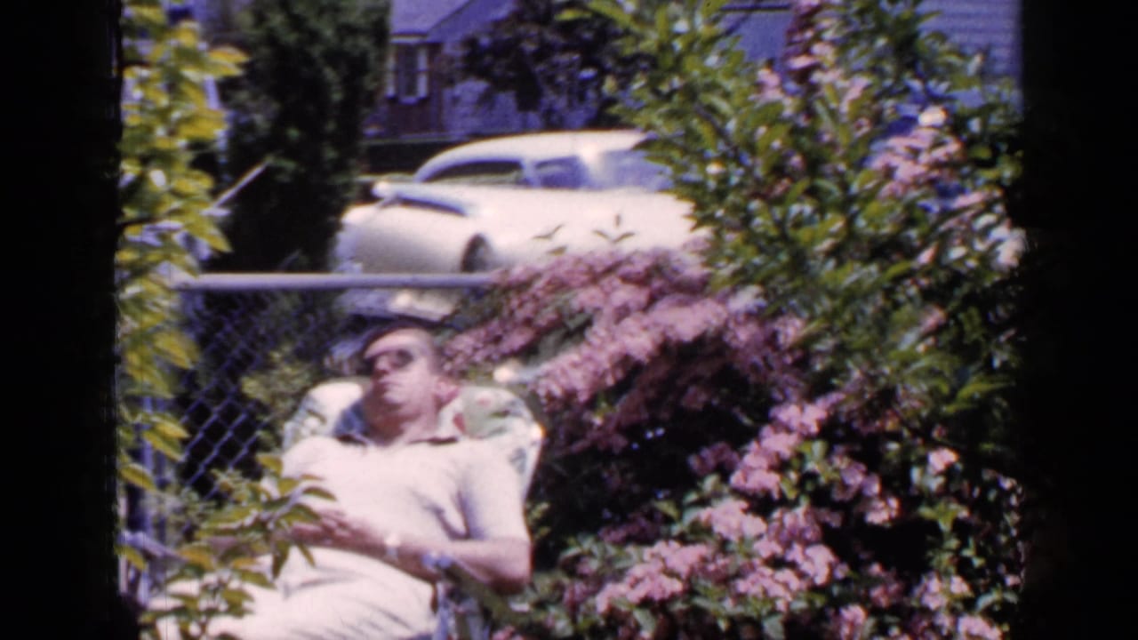 In 1965 Long Island home-movie footage, a man rests in a backyard lounge chair with flowering shrubs, fencing, and parked cars behind him.