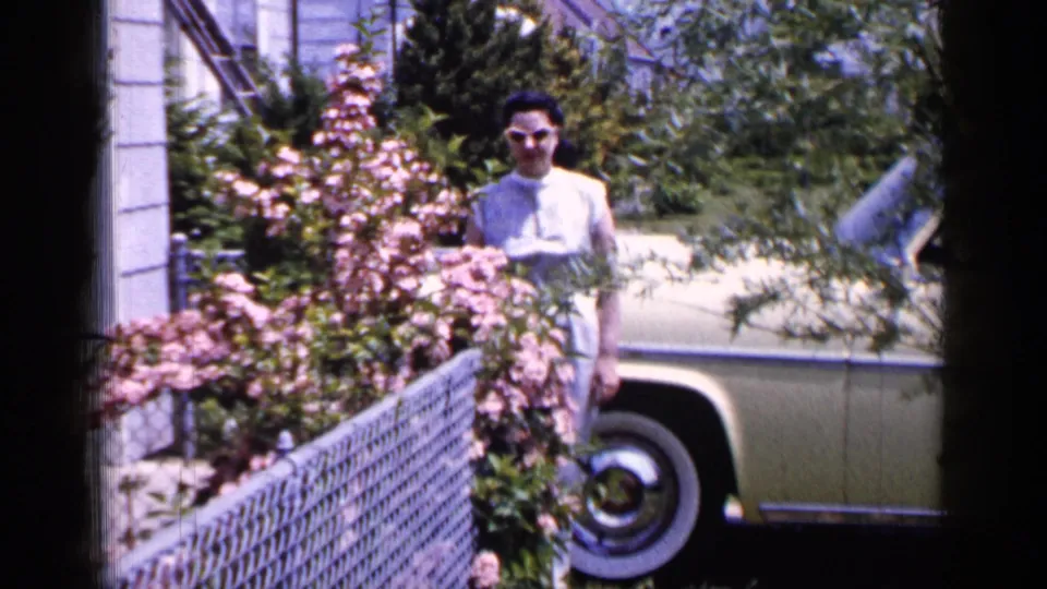 1965 Long Island frame of a woman in a light dress and glasses standing near a cream-colored vintage car, partly screened by flowering bushes and a chain-link fence.