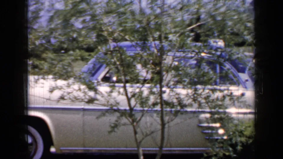 1965 Long Island home-movie frame showing a cream-colored mid-century sedan partly obscured by trees and a dark window edge.