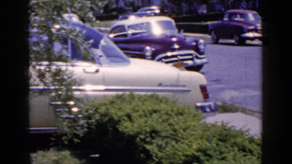 Home-movie view of a Long Island residential street in 1965, with classic cars, shrubs, and a cream sedan partly cut off by the frame.