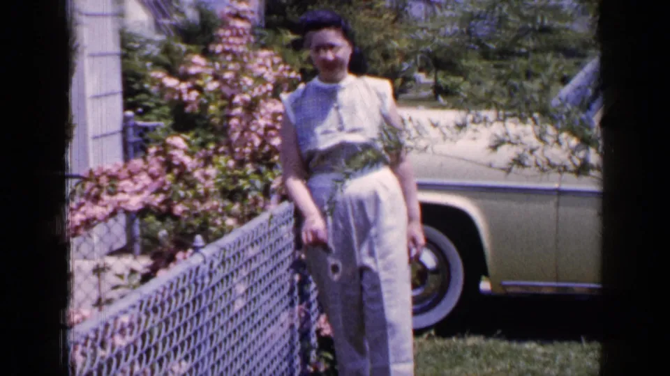 1965 Long Island home-movie frame showing a woman in a light dress beside a chain-link fence, pink flowers, lawn, and a parked sedan.