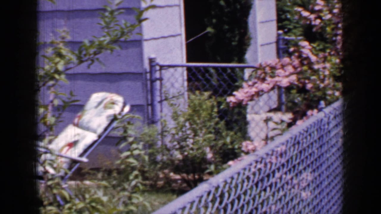 The camera moves across a Long Island residential yard, showing a folded lounge chair near shrubs, chain-link fencing, and light house siding.