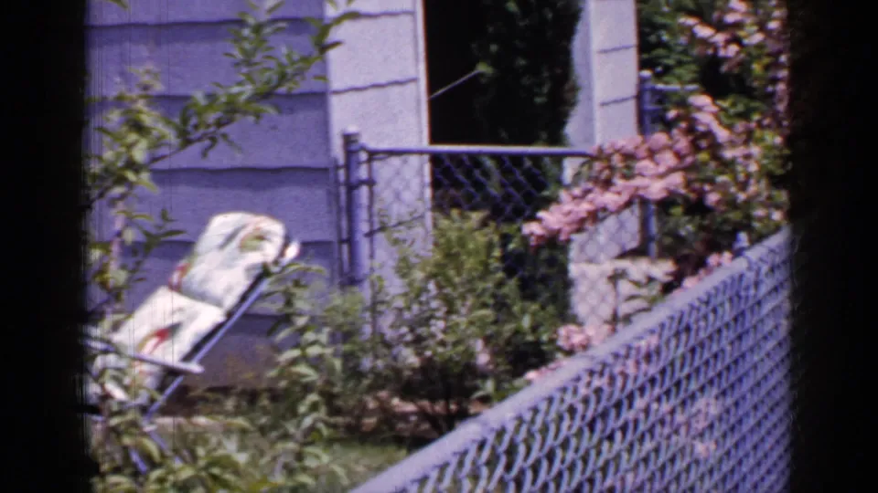 The camera moves across a Long Island residential yard, showing a folded lounge chair near shrubs, chain-link fencing, and light house siding.