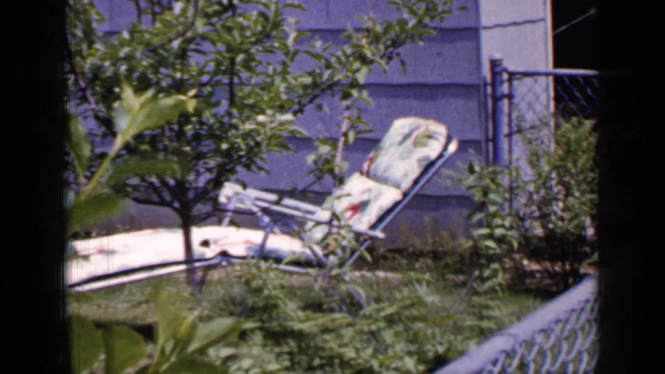 A metal lounge chair rests low in the grass near shrubs and young branches beside a light-sided Long Island house exterior.