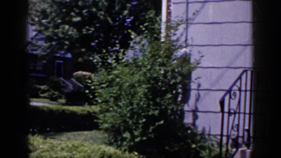The final frame settles on shrubs and lawn beside a light-sided Long Island house, with a dark porch area and metal railing at the edge.