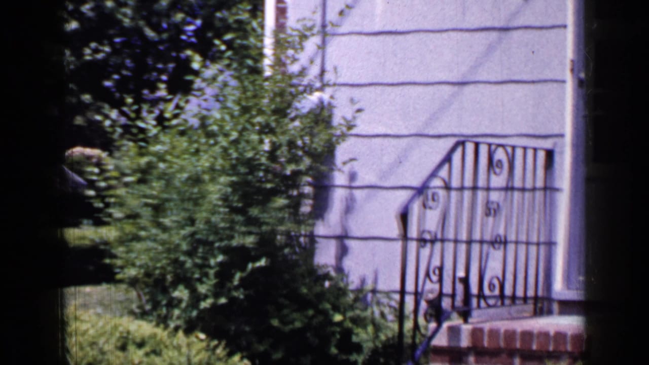 1965 Long Island home-movie frame showing a grey house facade with shrubs, lawn, front steps, and decorative black railing.