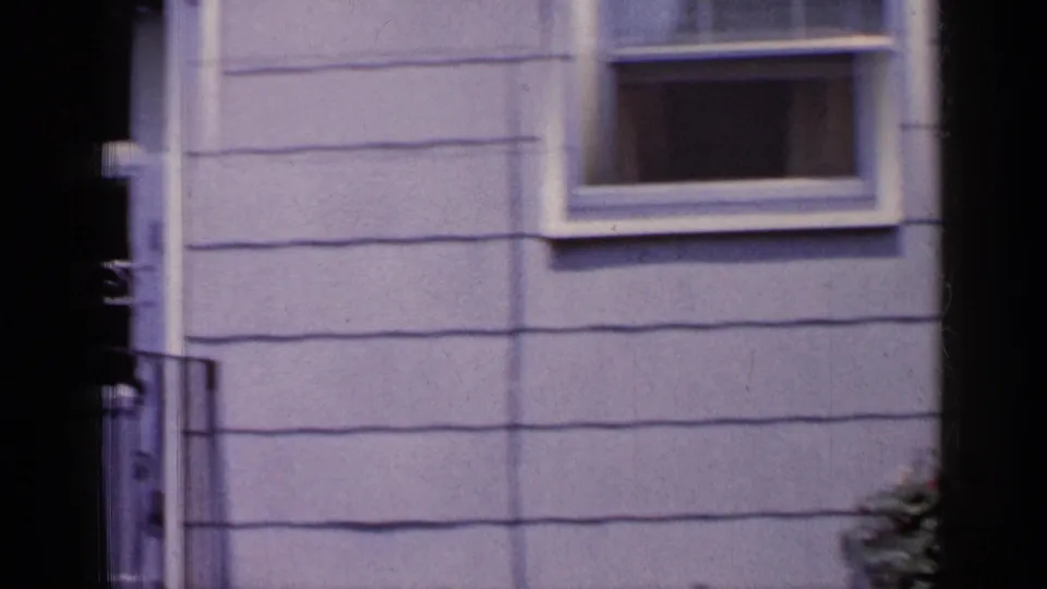 1965 Long Island home exterior frame showing grey horizontal siding, a small window, and part of a black railing near the lower edge.