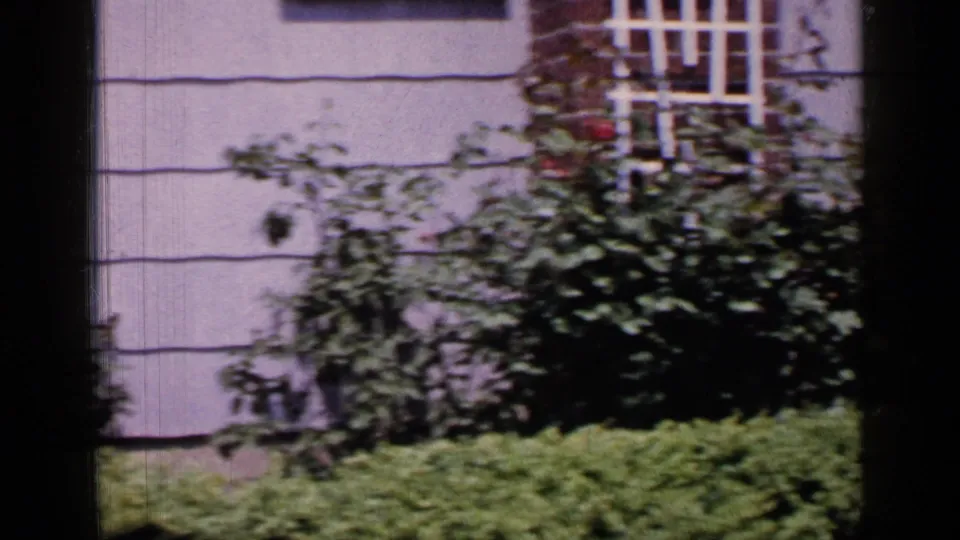 1965 Long Island home-movie frame showing a pale suburban house exterior, shuttered window, shrubs, hedge, and dark window-frame bars.