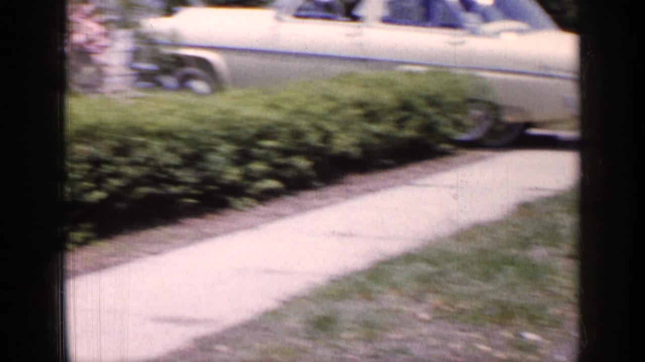1965 Long Island home-movie frame showing a sidewalk, grass, shrubs, and part of a parked car near a residential driveway.