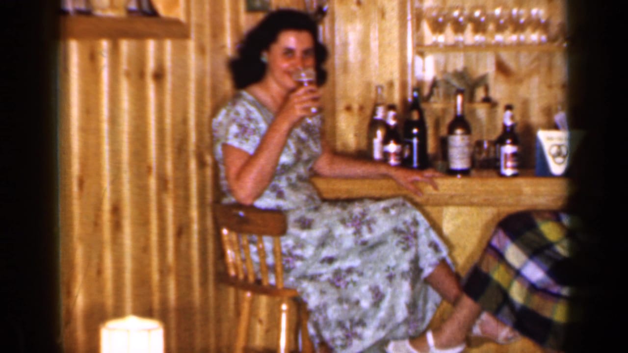 Woman in a floral dress seated at a wood-paneled bar with bottles and glasses.