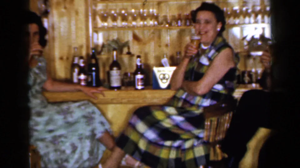 A woman in a plaid dress smiles while holding a drink at a wood-paneled home bar with bottles and glassware visible.