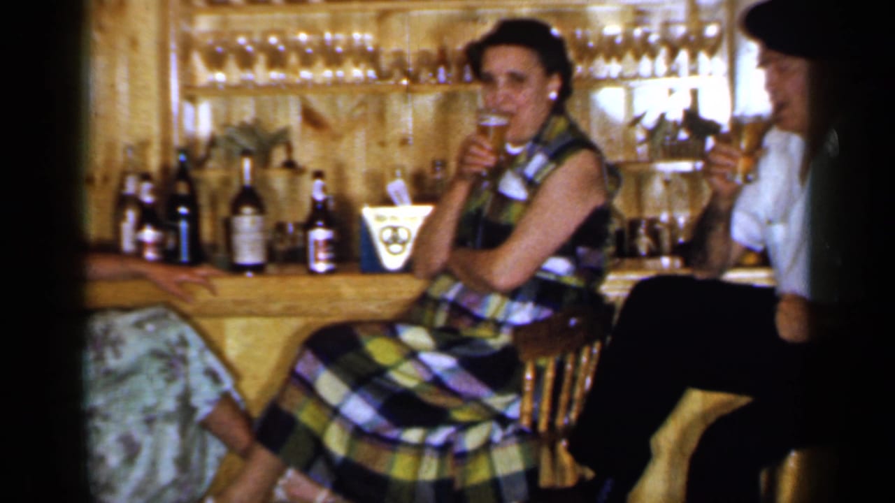 Two adults sit at a wooden bar with drinks and bottles behind them.