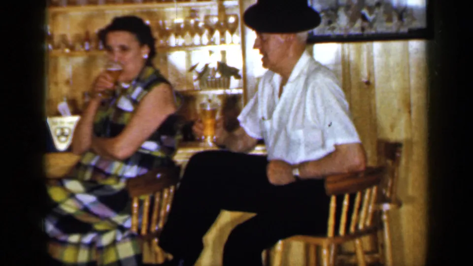 Home-movie frame from Long Island in 1965 showing two older adults seated at a rustic bar with glasses and bottles nearby.