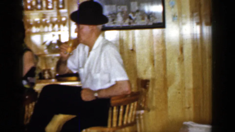 1965 Long Island home-movie still of a man in a white shirt and dark hat drinking at a wood-paneled bar.