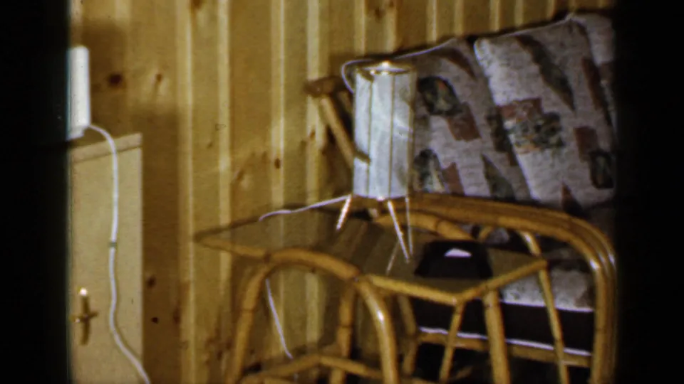 1965 Long Island home interior showing rattan seating, patterned cushions, a small lamp, side table, and wood-paneled wall.
