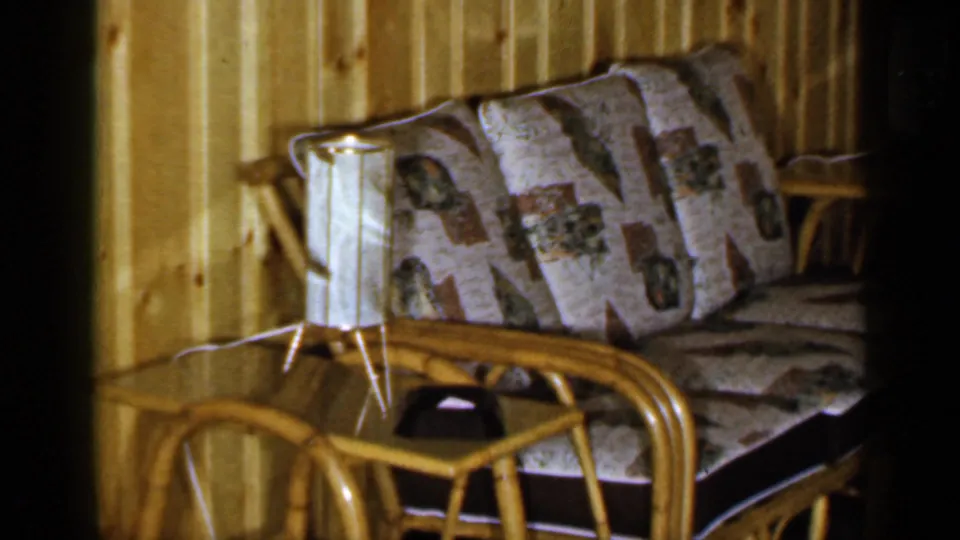 1965 Long Island frame with rattan sofa, patterned cushions, small table lamp, and wood-paneled domestic interior.