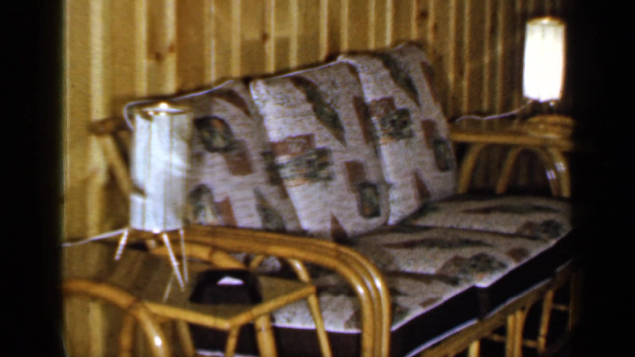 Rattan sofa with patterned cushions beside a glowing lamp in a wood-paneled room.
