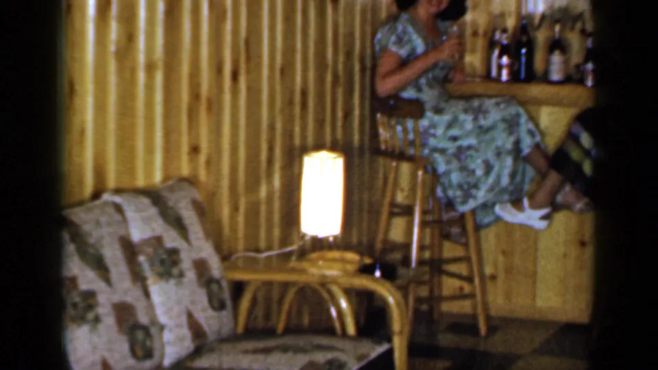 1965 Long Island home interior with rattan seating in the foreground and a woman partly visible on a stool at a small wood bar.